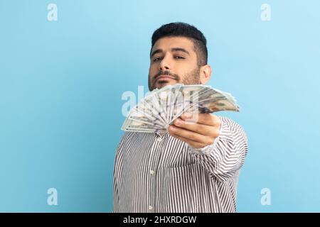 Ritratto di sicuro di sé ricco barbuto uomo d'affari che tiene fuori bollette del dollaro alla macchina fotografica, vantando i soldi vinti nella lotteria, indossando la camicia a righe. Studio interno girato isolato su sfondo blu. Foto Stock