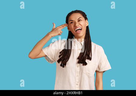 Ritratto della giovane donna depressa con ciamboli neri fa il gesto del suicidio, spara nel tempio, stoltisce intorno, indossando una camicia bianca. Studio interno girato isolato su sfondo blu. Foto Stock