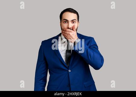 Ritratto di attraente businessman bearded chiusura bocca con mano, paura di dire troppo, imbarazzo, indossare abiti stile ufficiale. Studio interno girato isolato su sfondo grigio. Foto Stock