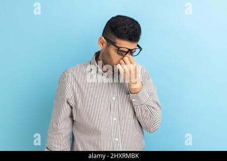 Ritratto di un uomo d'affari frustrato in occhiali che gli sfregano gli occhi, stanco di lunghe ore di lavoro sul laptop, dolore o dolore, indossando una camicia a righe. Studio interno girato isolato su sfondo blu. Foto Stock