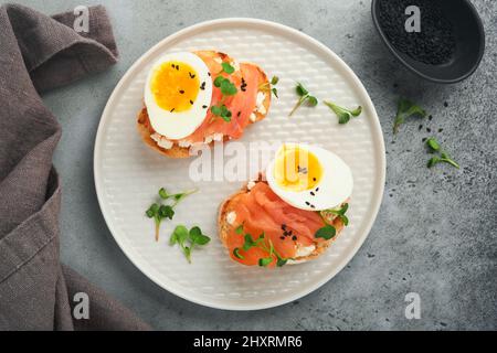 Sandwich con deliziosi condimenti salmone affumicato, uova, erbe e ravanello microgreens, semi di sesamo nero su piastra bianca su tavola grigia di cemento backgro Foto Stock