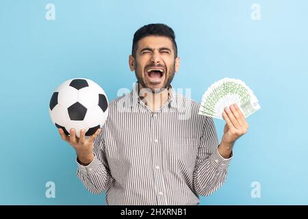 Estremamente felice uomo d'affari che tiene pallone da calcio e fan delle bollette euro, scommesse sportive, grande vittoria, gioire, urlare, indossare una camicia a righe. Studio interno girato isolato su sfondo blu. Foto Stock