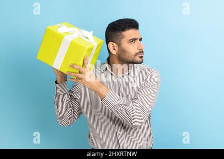 Ritratto di curioso businessman bearded che tiene e scuotendo avvolto scatola presente, essendo interessato che cosa all'interno, indossando camicia a righe. Studio interno girato isolato su sfondo blu. Foto Stock