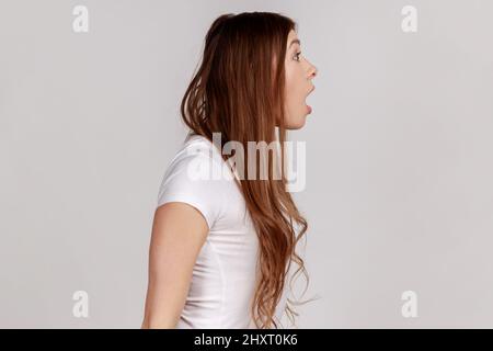 Ritratto della vista laterale di una donna stupita che guarda con la bocca aperta a sorpresa, espressione del viso scioccata, indossando una T-shirt bianca. Studio interno girato isolato su sfondo grigio. Foto Stock