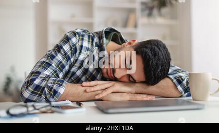 Giovane uomo esausto che dorme al lavoro seduto alla scrivania Foto Stock