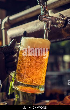 Primo piano verticale di un bicchiere di birra che viene riempito da un rubinetto contro uno sfondo sfocato Foto Stock