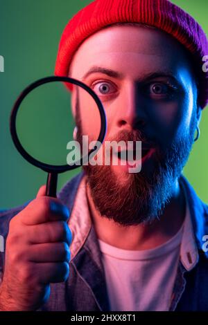 Ritratto di stupito hipster uomo in piedi, tenendo la lente d'ingrandimento e guardando la fotocamera, dopo aver stupito volto sorpreso. Studio interno girato isolato su colorato sfondo di luce al neon. Foto Stock