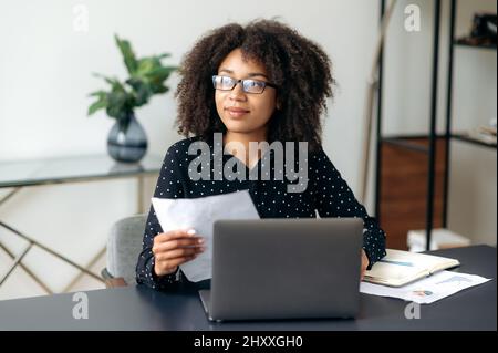 Attraente giovane adulto di successo, donna con pelle scura, leader aziendale, manager o lavoratore d'ufficio, si siede in ufficio, utilizza un notebook, lavora con documenti, grafici, guarda via, analizza le dinamiche economiche Foto Stock
