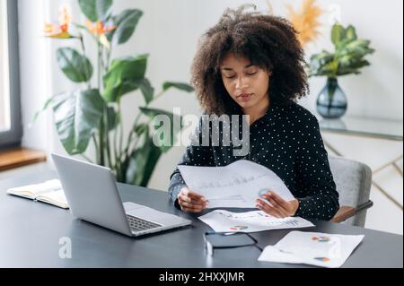 Donna focalizzata con pelle scura, freelance, manager, direttore aziendale, si siede in ufficio presso un laptop, lavora con i documenti in modo attento, analizza le dinamiche, sviluppa una strategia di lavoro, pianifica il business plan Foto Stock