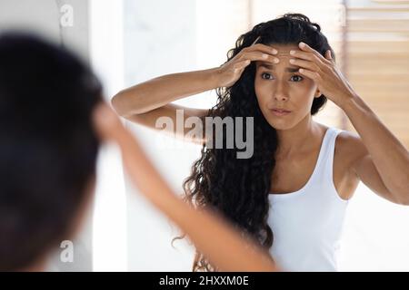 Giovane donna preoccupata che guarda nello specchio e che tocca le rughe sul suo volto Foto Stock