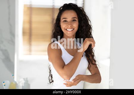 Bella giovane donna Apllying crema idratante sul gomito mentre in piedi in bagno Foto Stock