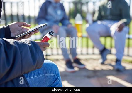 Primo piano di adolescenti con Mobile Phone Vaping e bere alcol nel Parco Foto Stock