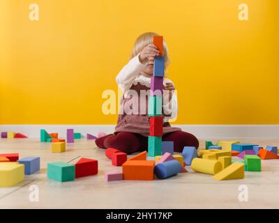 Una bambina è seduta sul pavimento in camera con pareti color giallo saturo e sta costruendo un'alta torre di blocchi multicolore di costruttore Foto Stock