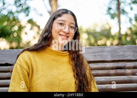 Giovane ragazza allegra con lunghi capelli scuri guardando la macchina fotografica con il sorriso mentre si siede su panca di legno nel parco su sfondo sfocato Foto Stock
