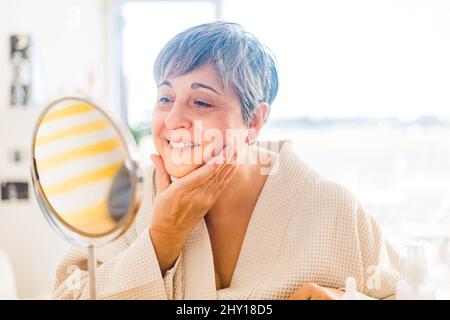 Donna adulta in accappatoio bianco che spalma crema idratante sul viso e guarda lo specchio Foto Stock