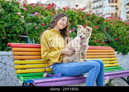 Giovane donna proprietario con capelli ricci e carino cane seduto su panca colorata vicino cespugli in fiore il giorno d'estate nel parco Foto Stock