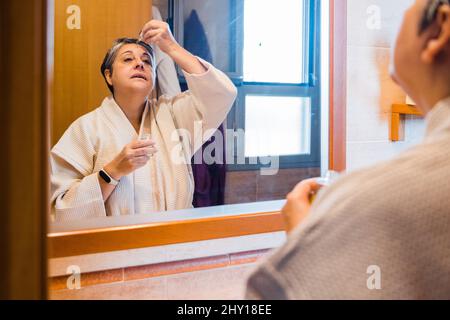 Donna di mezza età in accappatoio applicando siero idratante sul viso mentre si guarda nello specchio durante la routine di cura della pelle in bagno leggero Foto Stock