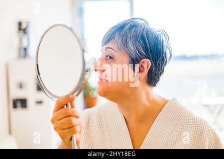 Contenuto femmina di mezza età con capelli corti in accappatoio bianco guardando lo specchio rotondo mentre si trova in piedi nella stanza della luce a casa Foto Stock
