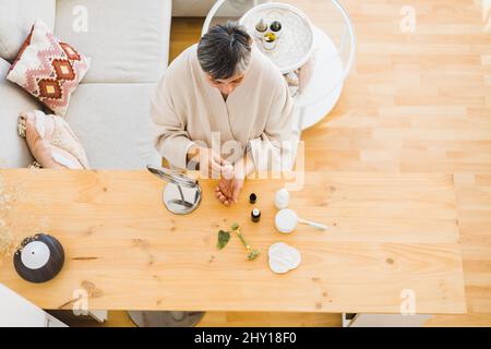 Da sopra della femmina di mezza età in accappatoio gocciolando siero a portata di mano mentre si siede a tavola durante la routine di cura della pelle a casa Foto Stock