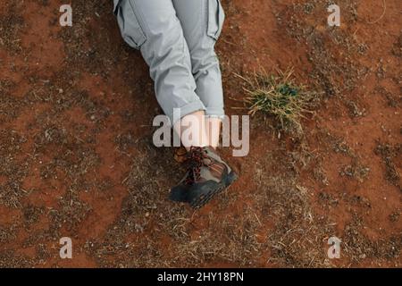 Da sopra, viaggiatore femminile non riconoscibile con scarpe da trekking adagiate su terreno asciutto durante il viaggio in terreno desertico Foto Stock