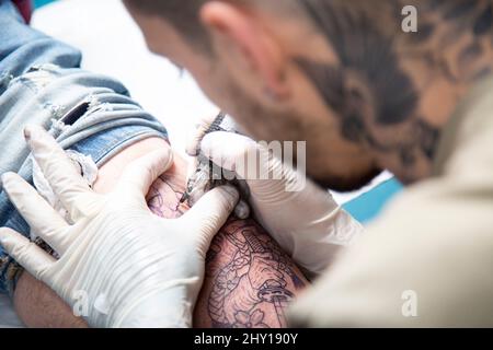 Da sopra raccolto senza volto professionale tatuaggio master in guanti in lattice facendo tatuaggio su gamba del cliente con moderna macchina rotativa tatuaggio penna Foto Stock