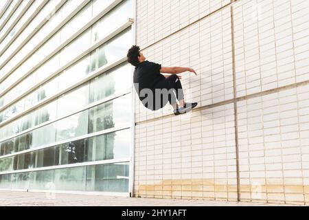 Vista laterale dell'uomo a tutta lunghezza in vestito nero che si esibisce in un parco acrobatico sulla parete dell'edificio Foto Stock