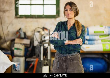 Calmo lavoratore femminile guardando la macchina fotografica con le braccia incrociate e faccia seria mentre si trova in azienda agricola con varie forniture durante il lavoro Foto Stock