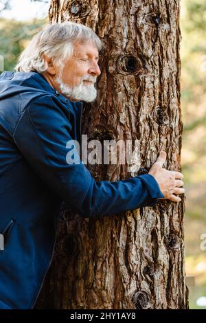 Vista laterale dell'uomo anziano con capelli grigi e barba che abbraccia l'albero nella foresta autunnale con occhi chiusi su sfondo sfocato Foto Stock