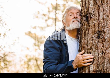 Uomo anziano con capelli grigi e barba che abbraccia l'albero nella foresta autunnale con occhi chiusi su sfondo sfocato Foto Stock