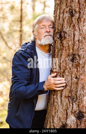 Uomo anziano con capelli grigi e barba che abbraccia l'albero nella foresta autunnale con occhi chiusi su sfondo sfocato Foto Stock