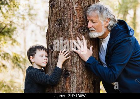 Uomo anziano con capelli grigi e barba e carino ragazzo abbraccio albero in autunno foresta con occhi chiusi su sfondo sfocato Foto Stock