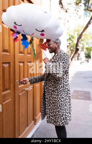 Vista laterale di una femmina afro-americana positiva con palloncino nuvoloso che apre la porta d'ingresso con la chiave mentre si trova sulla strada Foto Stock