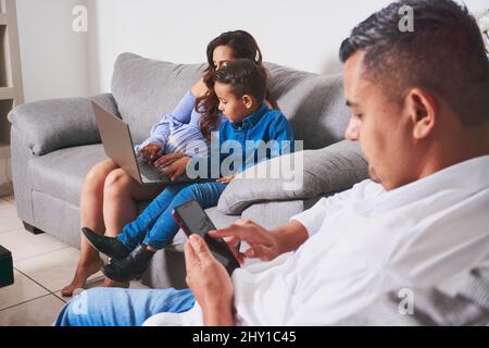 Famiglia ispanica con ragazzo surf smartphone moderno e computer portatile, mentre trascorrere il tempo insieme su un comodo divano in un soggiorno leggero Foto Stock