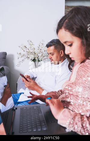 Vista laterale della ragazza ispanica che batte sul netbook mentre si siede sul comodo divano vicino al padre che naviga smartphone in salotto Foto Stock