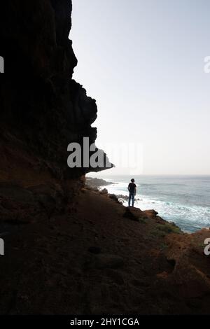 Vista posteriore dell'irriconoscibile viaggiatore maschile in piedi sul bordo di ripida scogliera e osservando il mare sotto il cielo blu sulla costa dell'isola di Lanzarote Foto Stock