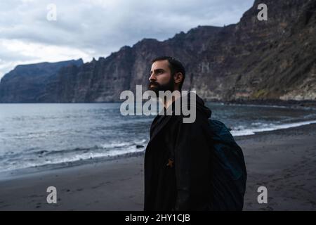 Vista laterale del turista maschio barbato in outerwear con zaino in piedi su una spiaggia sabbiosa bagnata dall'oceano contro ripide scogliere rocciose e ammirare la natura du Foto Stock