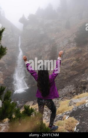 Vista posteriore del corpo del viaggiatore anonimo femminile con braccia sollevate in piedi su un pendio roccioso mentre si ammira la cascata in Highlands Foto Stock