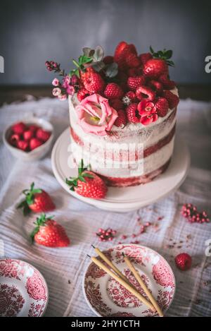 Deliziosa torta di velluto rosso sormontata da fragole fresche e decorata con fiori servita al tavolo con tovaglia bianca in cucina Foto Stock