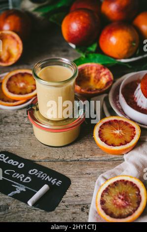 Vasetti di vetro con dolce e saporita cagliata d'arancia rossa vicino a un mucchio di citruse rosse poste su un tavolo di legno con centrifuga Foto Stock