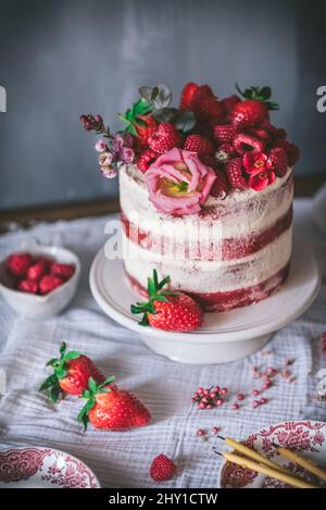 Deliziosa torta di velluto rosso sormontata da fragole fresche e decorata con fiori servita al tavolo con tovaglia bianca in cucina Foto Stock