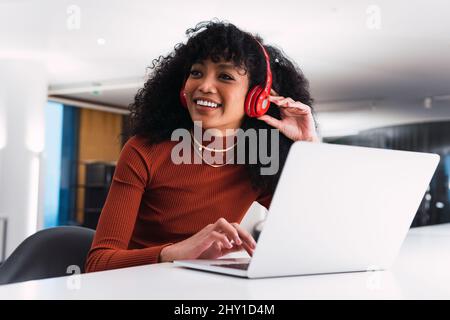 Contenuti afroamericana femminile in cuffie utilizzando il telefono seduto al tavolo con il portatile in un ambiente di lavoro moderno Foto Stock