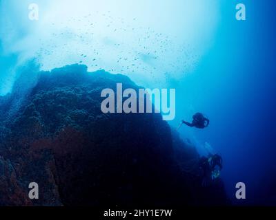 Subacquei professionisti irriconoscibili in mute e pinne che nuotano in prossimità di formazioni rocciose irregolari con superficie irregolare in mare blu profondo con acqua limpida Foto Stock