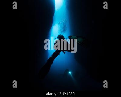 Subacquei professionisti irriconoscibili con scuba e pinne che nuotano in una stretta grotta scura con formazioni rocciose al di sotto del mare in acque profonde e limpide Foto Stock