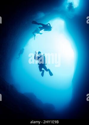 Subacquei professionisti irriconoscibili con scuba e pinne che nuotano in una stretta grotta scura con formazioni rocciose al di sotto del mare in acque profonde e limpide Foto Stock