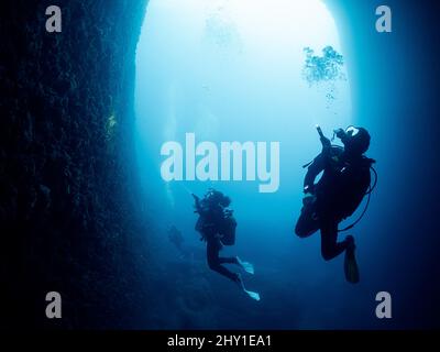 Subacquei professionisti irriconoscibili con scuba e pinne che nuotano in una stretta grotta scura con formazioni rocciose al di sotto del mare in acque profonde e limpide Foto Stock