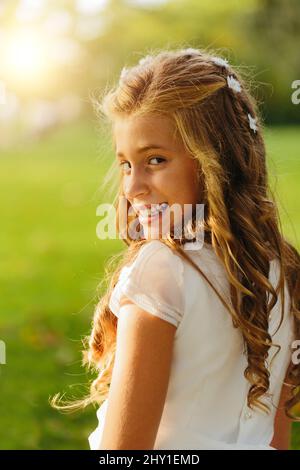 Felice giovane femmina in prima comunione vestito e collana sorridente e guardando la macchina fotografica mentre riposa in verde prato il giorno di sole Foto Stock