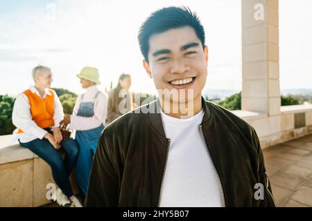 Giovane uomo asiatico sicuro che sorride alla macchina fotografica mentre si è in piedi con gli amici all'aperto Foto Stock