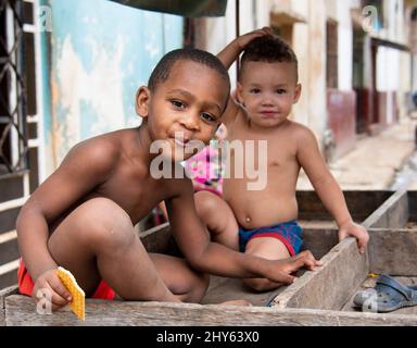 Il giovane ragazzo cubano mangia un biscotto mentre il suo amico guarda e sorride la macchina fotografica mentre si siede su un carrello in un quartiere a l'Avana, Cuba. Foto Stock