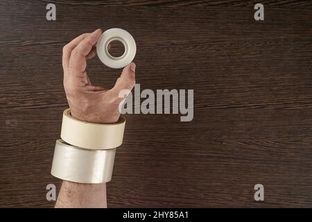 Vista dall'alto della mano dell'uomo che manica diversi tipi di nastri adesivi con fondo marrone Foto Stock
