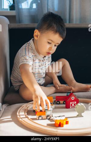 Ragazzino caucasico che gioca con il treno di legno mentre si siede sul pavimento, costruendo la ferrovia giocattolo a casa. Foto Stock